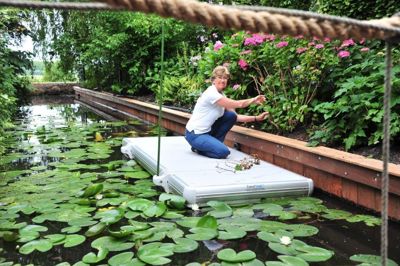 Kvinna sitter på huk på en EasyFloat arbetsflotte och beskär växter i en damm med näckrosor, smidigt för arbete på vatten där det är svårt att komma åt